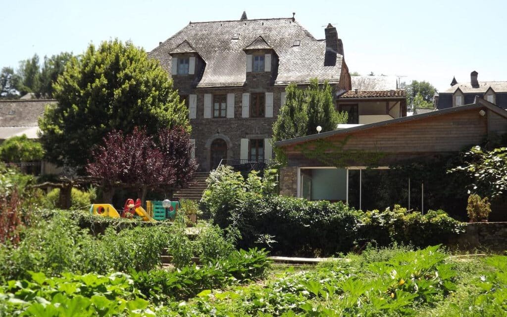 Jardin luxuriant de l'Hostellerie des Lauriers, avec une variété de plantes vertes et un petit espace de jeux pour enfants, offrant une oasis naturelle devant l'architecture traditionnelle en pierre de l'hôtel.