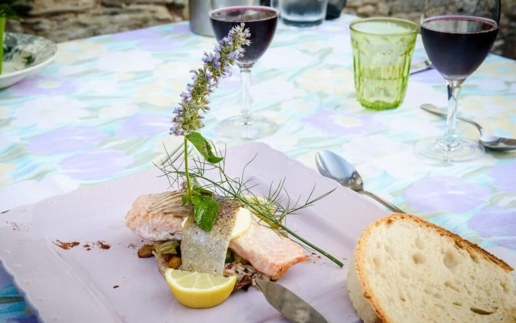 Plat gastronomique de l'Hostellerie des Lauriers présentant un filet de poisson délicatement assaisonné accompagné de citron et d'herbes fraîches, servi avec du pain croustillant et un verre de vin rouge, sur une table dressée avec une nappe colorée.