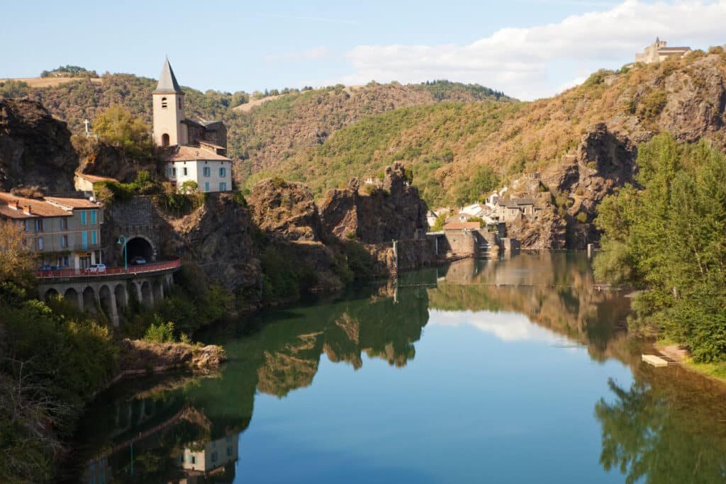 Le pittoresque village d'Ambialet niché dans un méandre de la rivière Tarn, avec son église perchée sur une colline escarpée et son reflet miroitant dans l'eau calme, offrant un cadre paisible et historique.