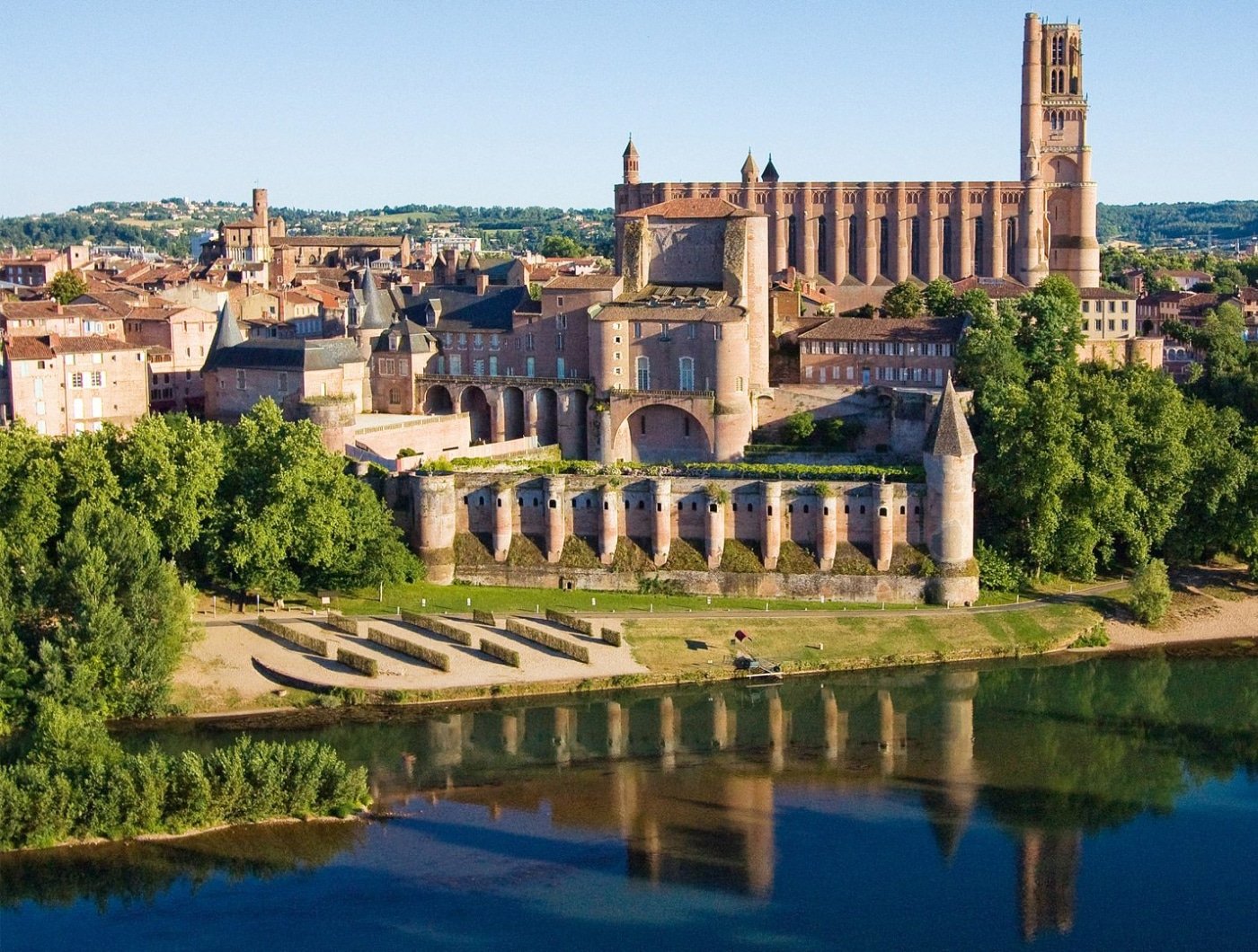 La Cité épiscopale d'Albi se reflétant dans les eaux tranquilles du Tarn, avec sa majestueuse cathédrale Sainte-Cécile et son palais fortifié, entourés de bâtiments historiques ocre, sous un ciel bleu limpide.